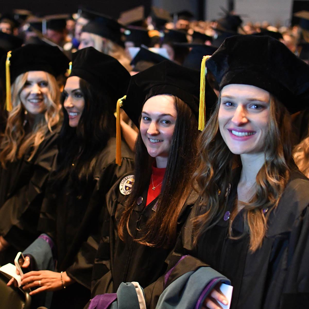 Students in graduation regalia are seated at the ceremony, looking to the camera and smiling